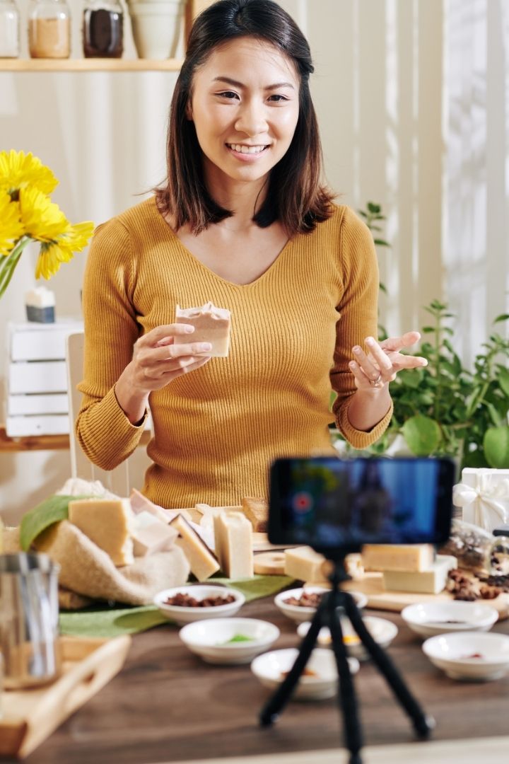 woman standing at counter, holding soap, filming on phone on mini tripod